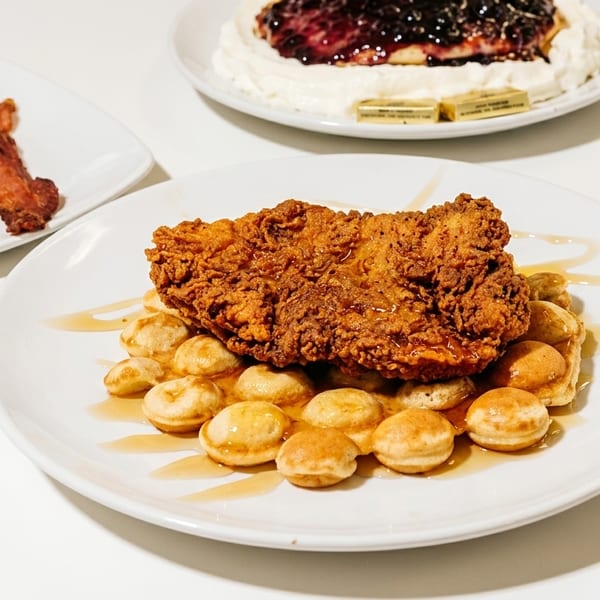 Crispy fried chicken and mini waffles drizzled with syrup, with bacon and berry pancakes in the background.